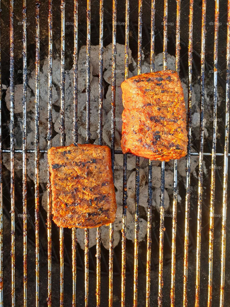 a top down portrait of two steaks grilling on a barbecue grill during a hot summer day.