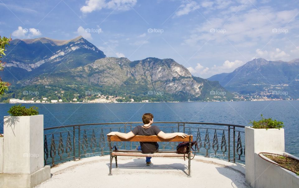 Homem branco sentado em banco sob a forte luz do sol, a observar a linda paisagem de Lagos e montes na cidade de Bellagio, Itália.