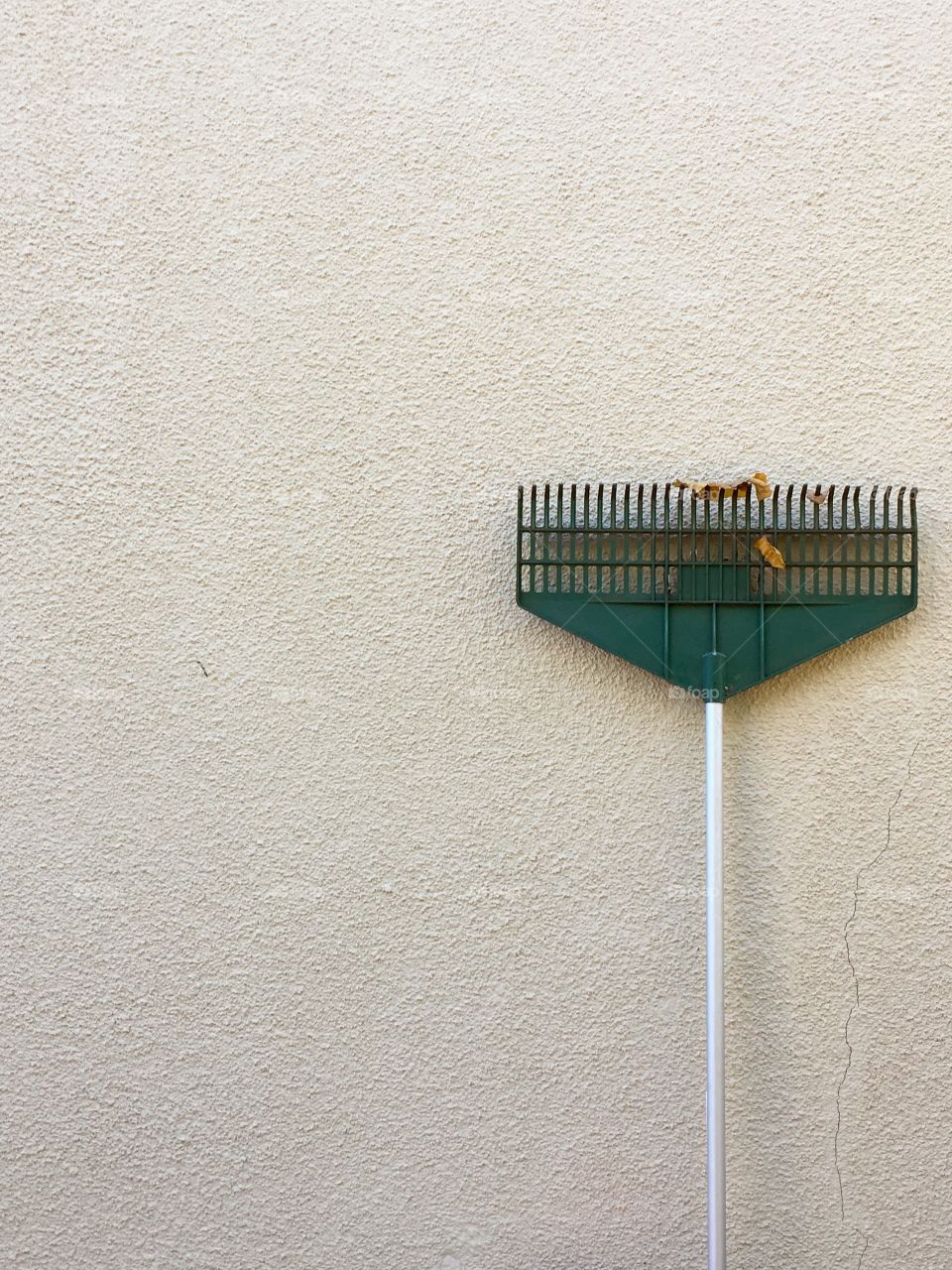 Green garden rake leaning up against stucco exterior wall, minimalist negative space