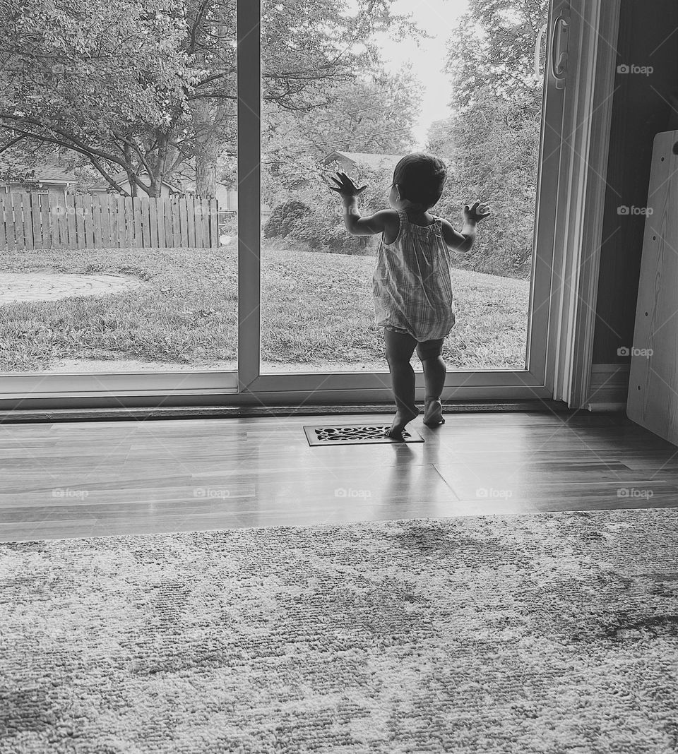 Black and white portrait of a baby looking outside, baby with hands in glass peering outside, baby looks at backyard of home, curious baby peers out window 