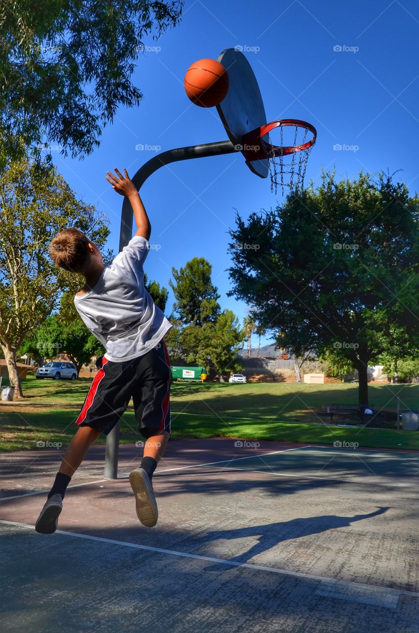 The boy is learning to shoot the basketball from one side. The pic is made while he is in mid-air, shooting. The ball is on the right direction. Will it go in?