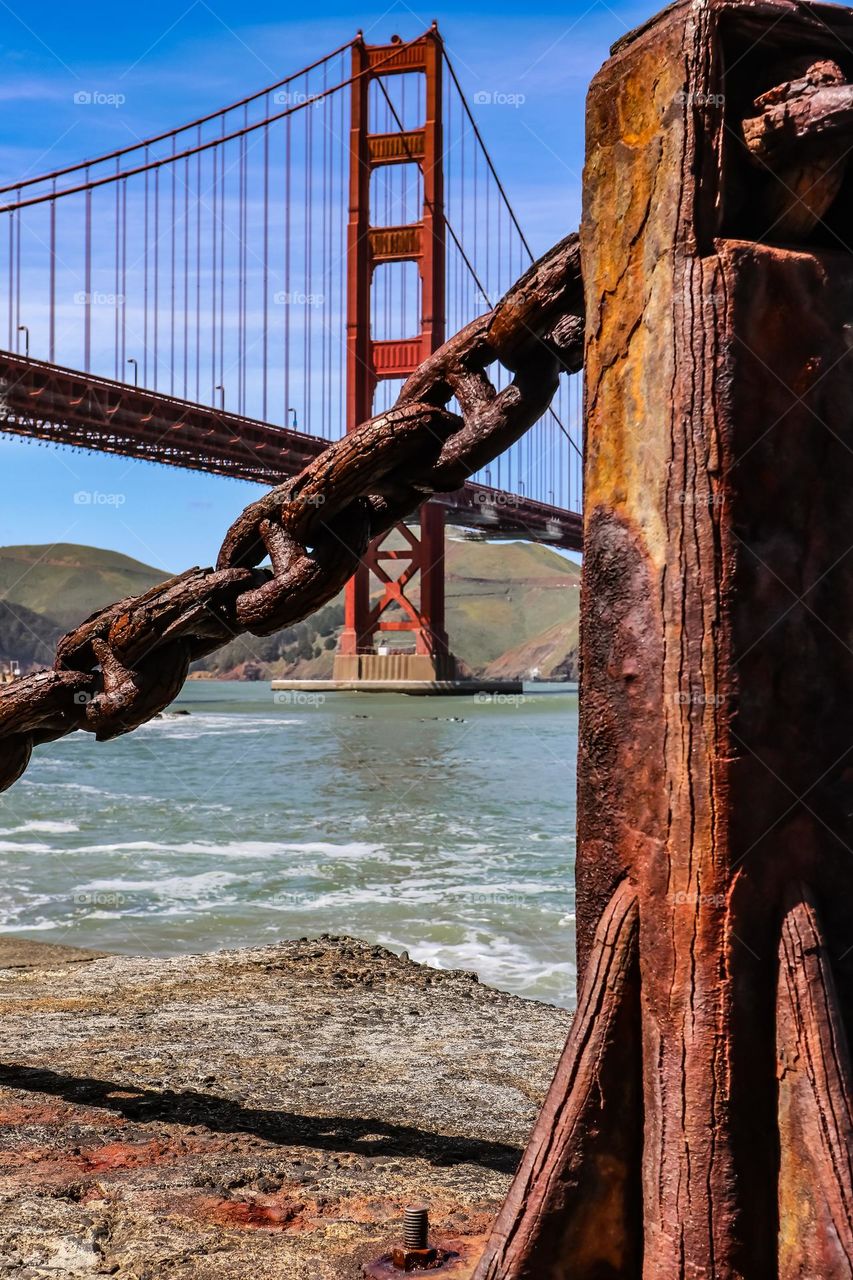 Ground level view of the Golden Gate Bridge from Fort Point in San Francisco California through the rusted iron chain link guardrails and posts that have oxidized over time appearing almost wood grain like.