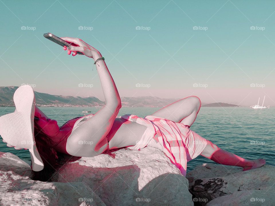 A young woman lying down on the rock by the sea and enjoys taking a photo of herself