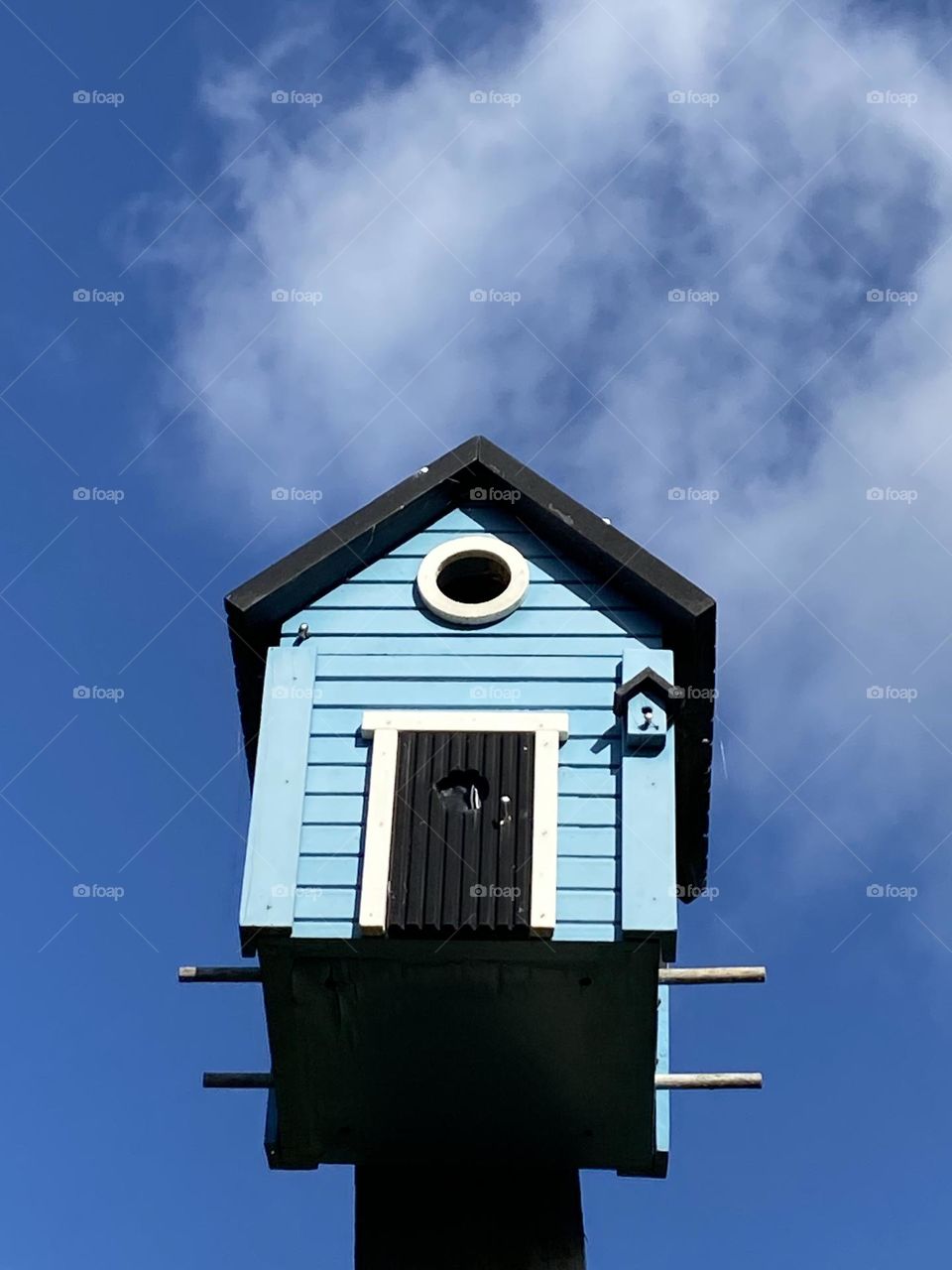 A blue and white birdhouse, photographed from bottom to top, juts into the deep blue summer sky. Some small clouds in the sky give the image additional character. 