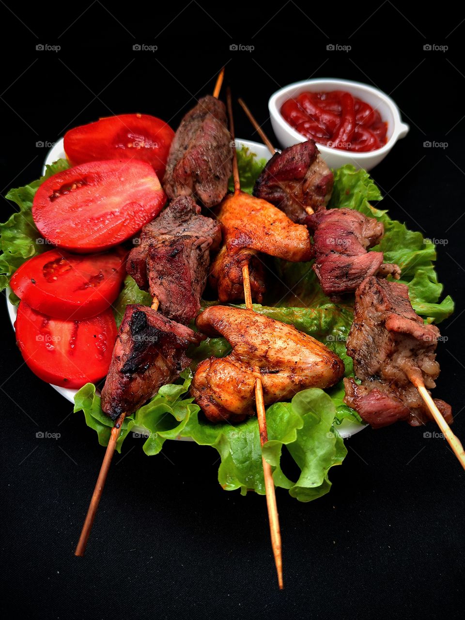 A white plate on which lie: green lettuce leaves, pieces of fried meat on a skewer, roasted wings on a skewer, chopped red tomatoes. In the background is a white gravy boat with red ketchup. Black background