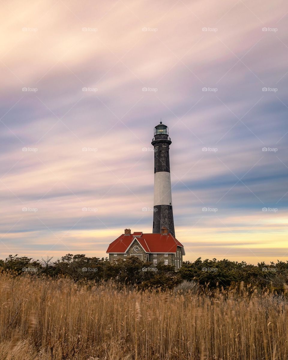 Long wispy clouds catching golden light from a sunset streaking over a tall lighthouse.   