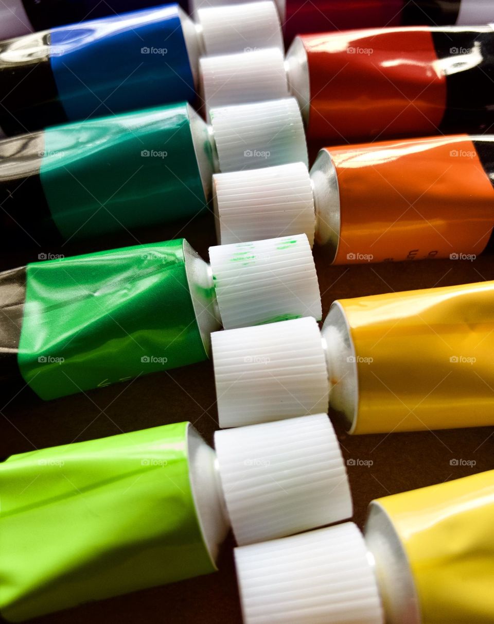  Closeup row of colorful oil paint tubes. Focus is on the white plastic caps contrasting with rich colors in green and orange tones.