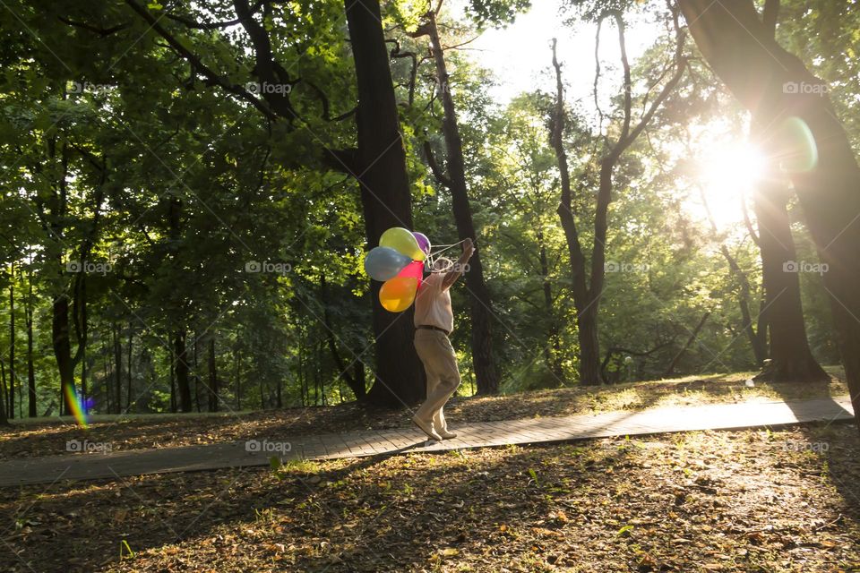 A lonely pensioner walks along the paths of the park on his birthday with balloons against the backdrop of the setting sun and trees.