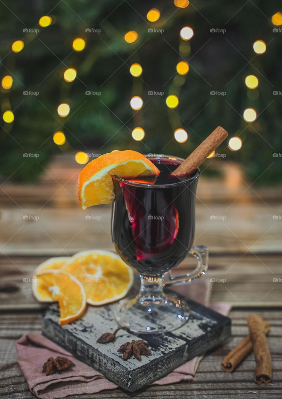 One glass of hot traditional drink mulled wine with cinnamon, anise and orange slices on a cutting board, stands on a wooden table with blurred ion, spruce branches, burning garland and bokeh, vtd side close-up. Traditional