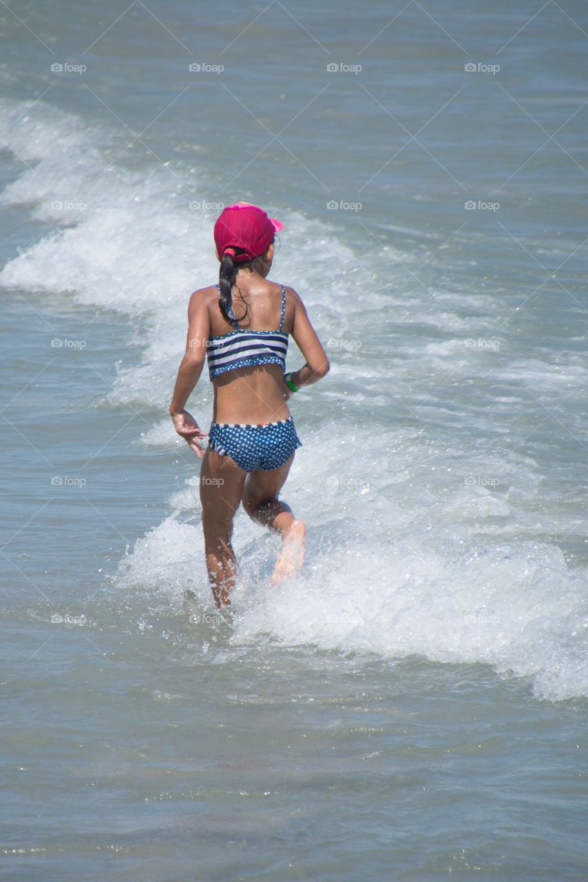 A girl protecting her head with a cap against the burning sun in spain runs through the waves of the ocean