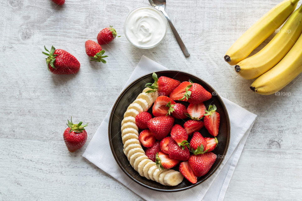 Top view to the strawberries and bananas salad on the plate at table near the strawberry fruits and bowl with yogurt.