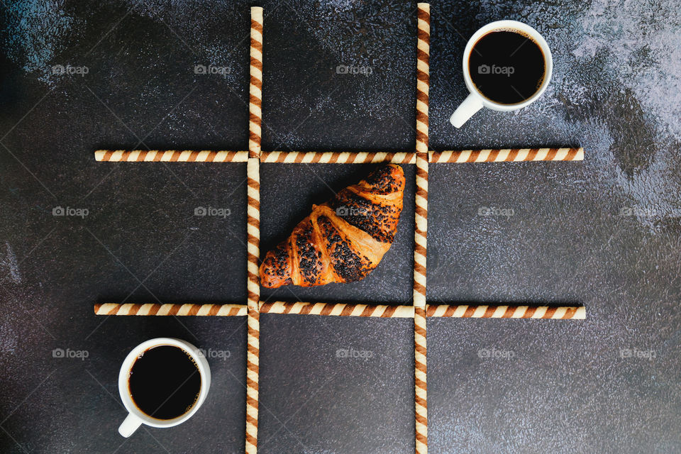 Working morning with cups of hot black coffee and sweet croissant on black background. Tic tac toe game. Top view. Flat lay. Creative concept of modern breakfast food.