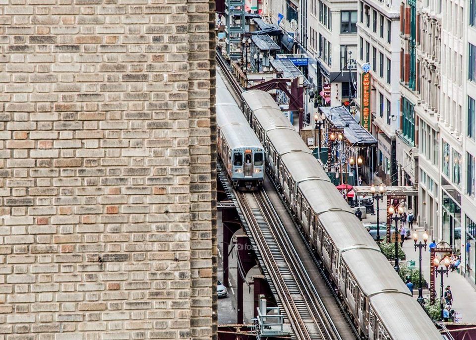 View from above of two trains arriving into the train station, surrounded by city buildings , downtown Chicago area. Underneath cars in the traffic and people walking down the street along the shops and restaurant 