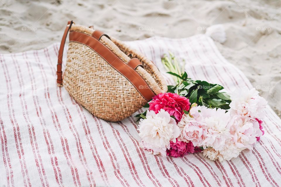 Wicker bag and bouquet of pink and white peonies on blanket on the sand, picnic on the beach