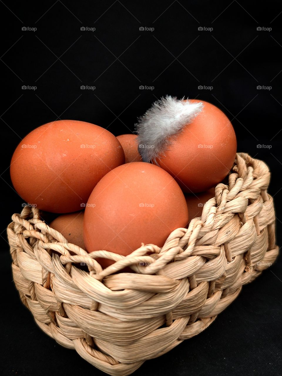 a wicker basket made of wood with brown chicken eggs in it.  on one egg a chicken feather.  Black background