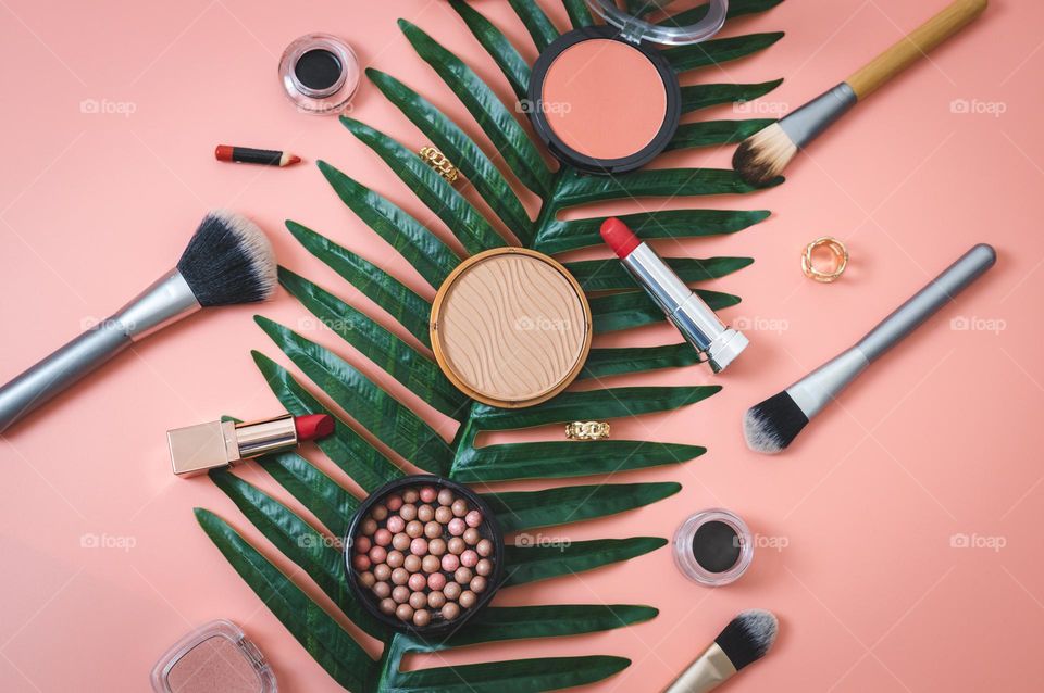 Makeup set of foundation granular face powder,brushes,brushes,eye pencil,red lipstick,soft black eye shadow on a palm tree lying diagonally on a pink background,flat lay close-up. Concept female cosmetics, beauty salon.