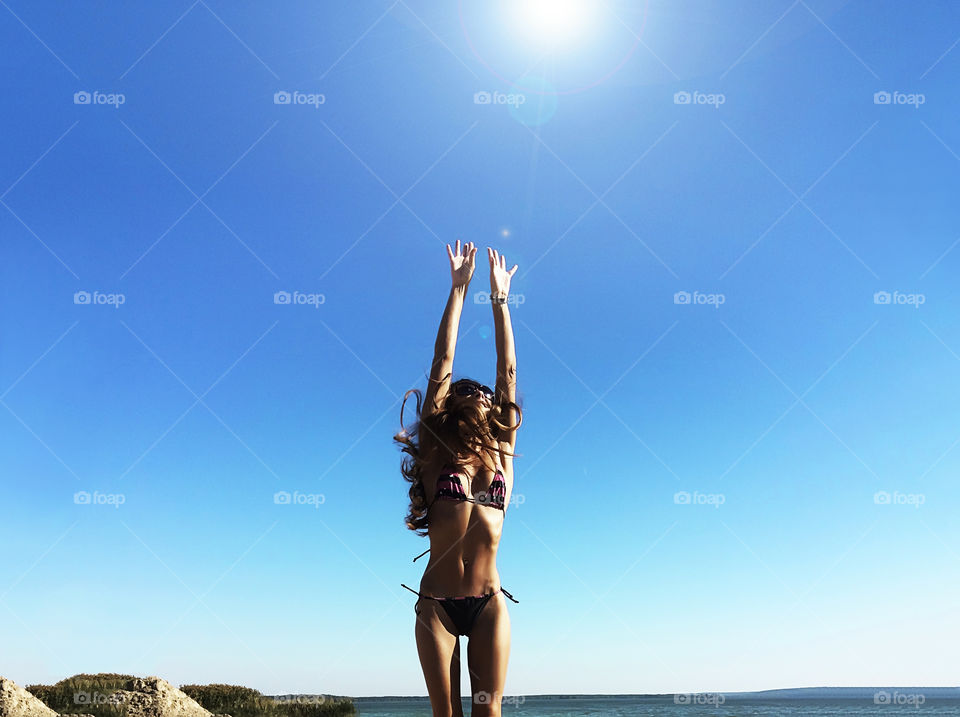 Young woman with long hair happily jumping towards bright summer sun on blue sky background 
