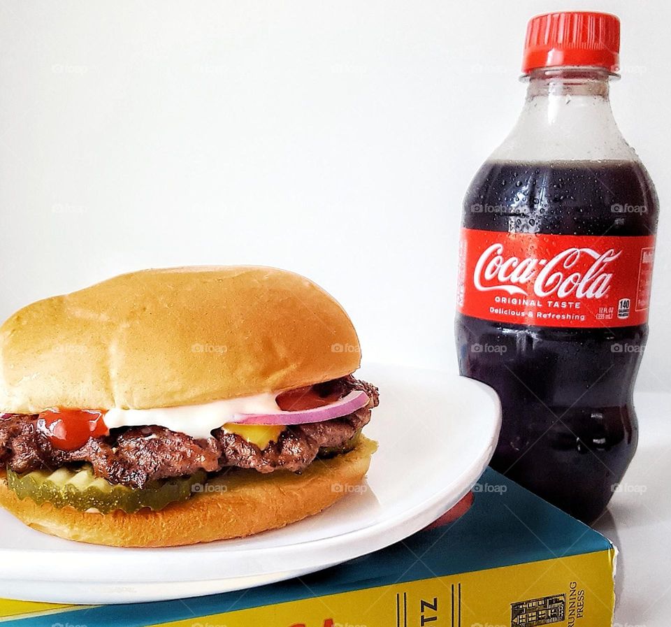 Cheeseburger with ketchup mayonnaise red onion pickle chips and American cheese on a white plate placed on top of a book next to a cold Coca-Cola soda pop