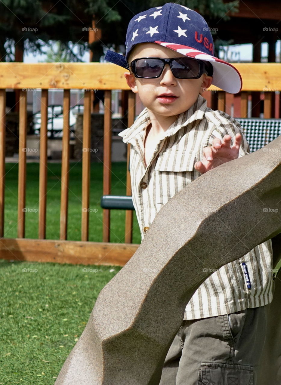 A young boy in a playground structure wearing his hat cocked and sunglasses on a sunny summer day. 