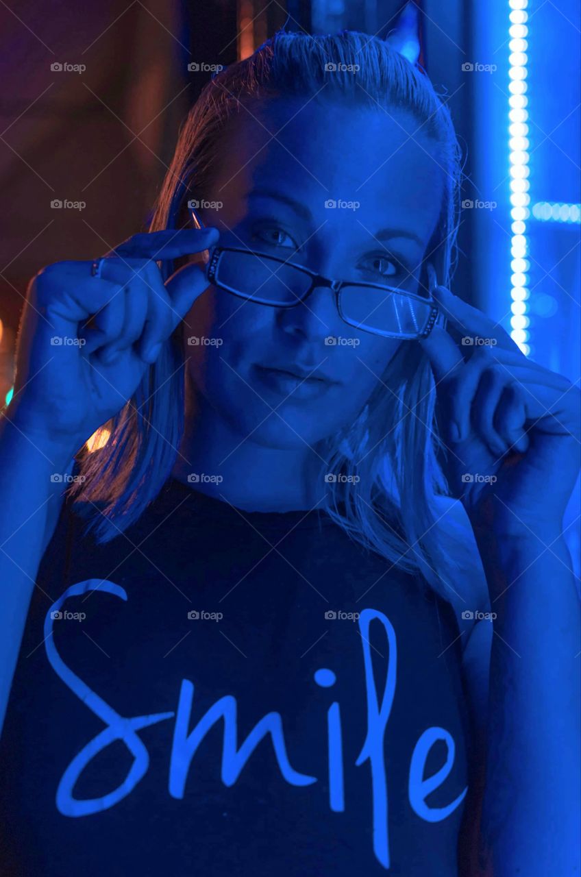 image of girl standing in blue window light wearing a t-shirt that says "smile" while tilting her glasses forward and looking into the camera