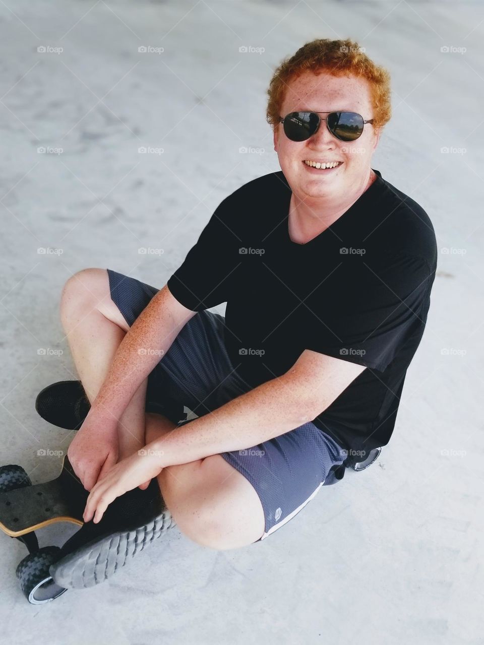 Man sitting on his skateboard wearing sunglasses & smiling