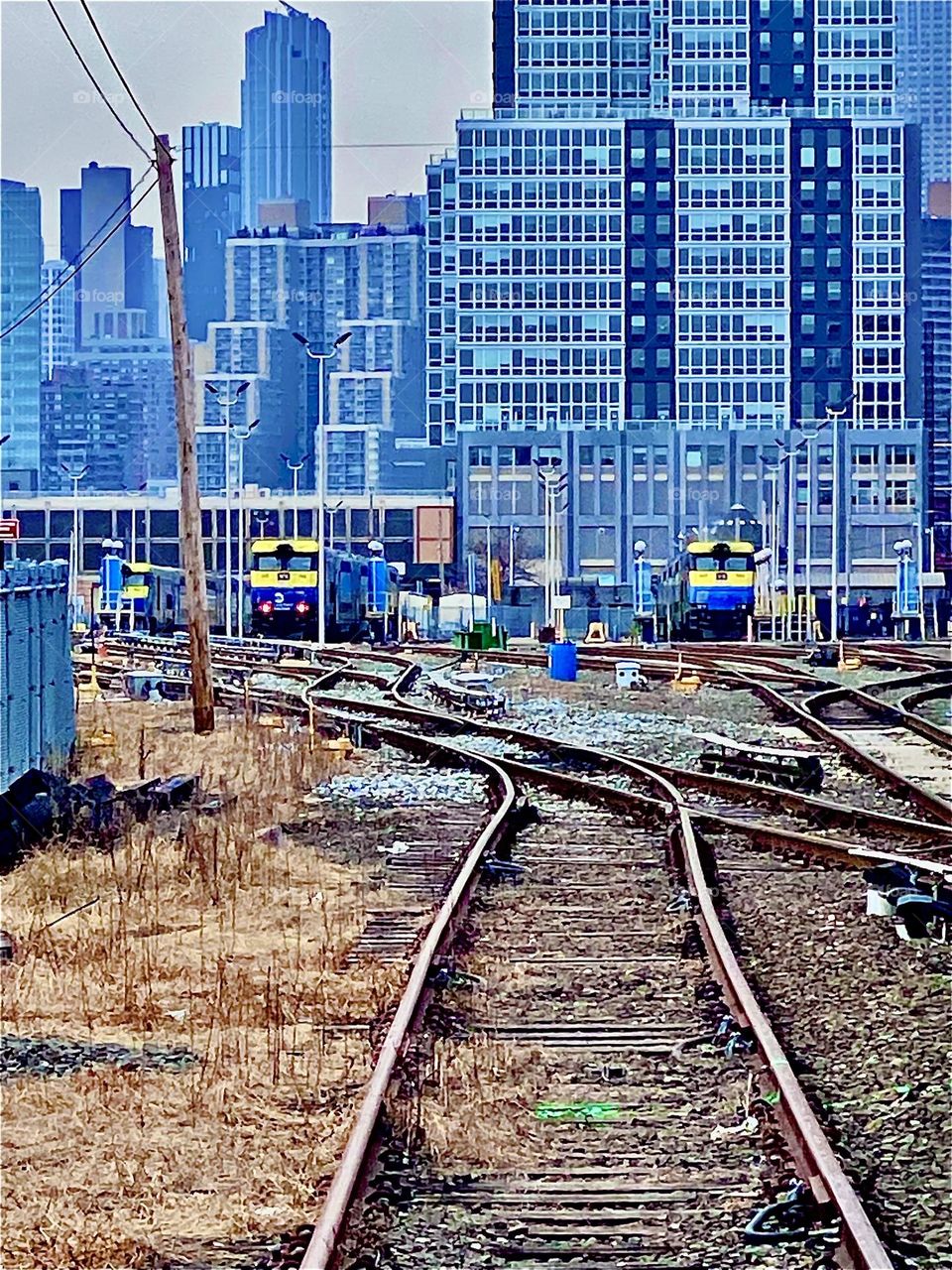 Train tracks and bright yellow and blue trains approaching on MTA grounds one crosses over regularly to ascend the pedestrian way onto the Pulaski Bridge for example using the staircase. 2021. Hypnotic Productions