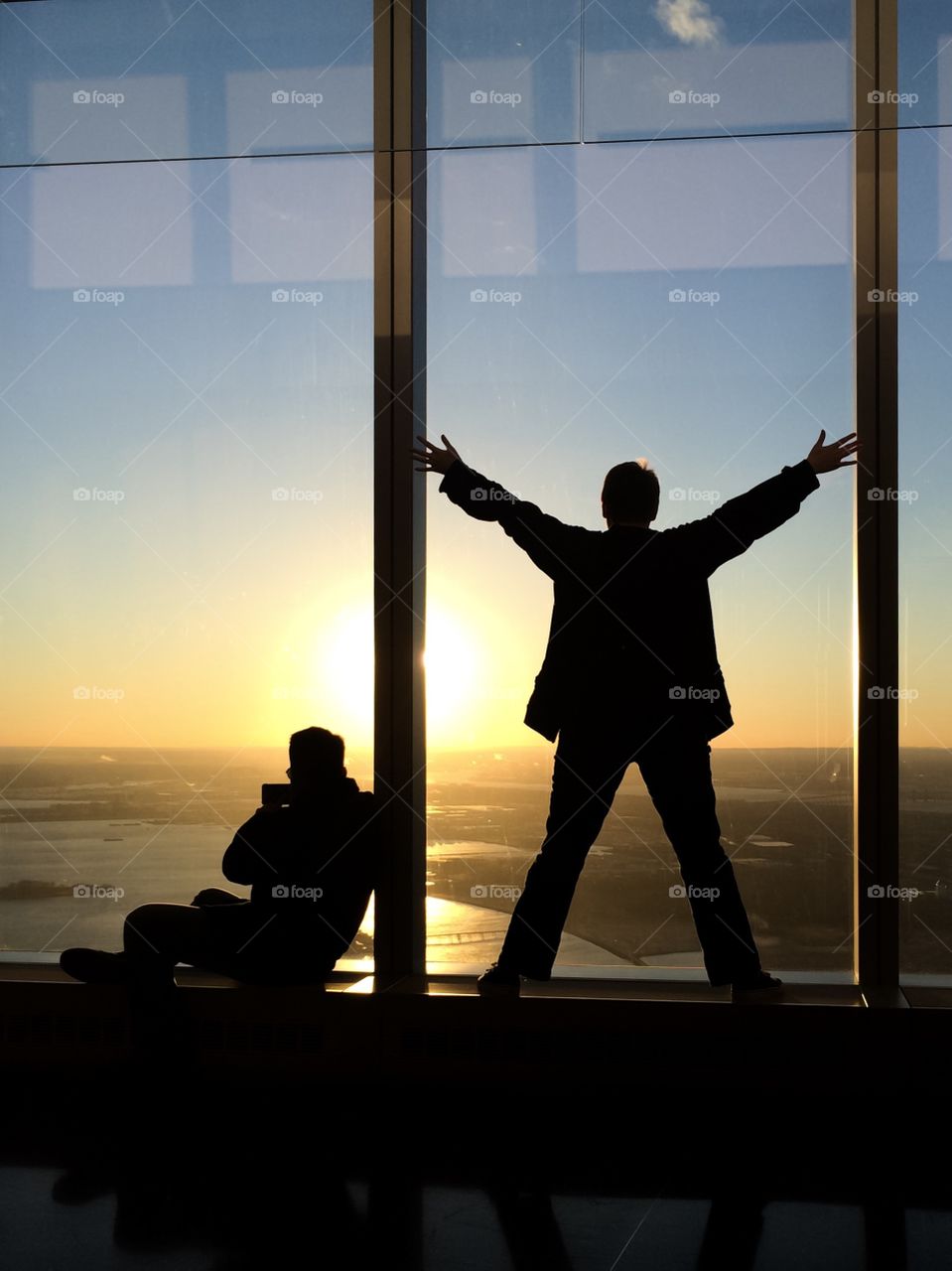 Two people watching the sun go down from the viewing gallery of a skyscraper. One person is taking a picture with their phone and one person is enjoying the moment