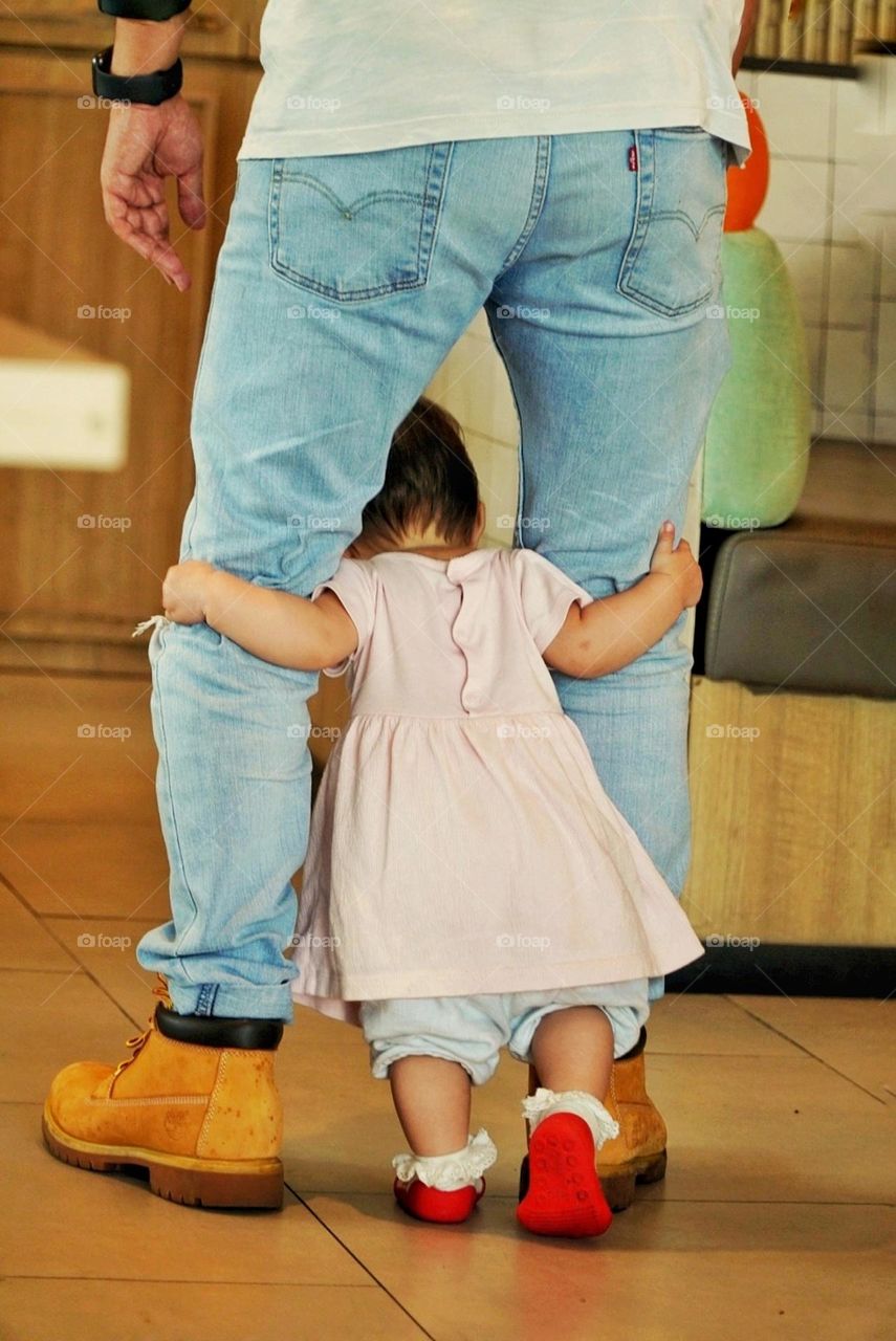 a two-year-old baby who is learning to walk holding on to his father's knee