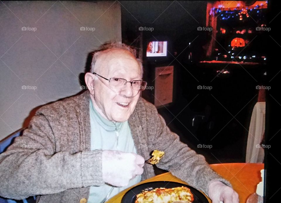 Older man, Grandfather, eating a plate of homemade lasagna. He's smiling, happy, wearing a sweater, eyeglasses and is using oxygen to help him breathe with a tube.