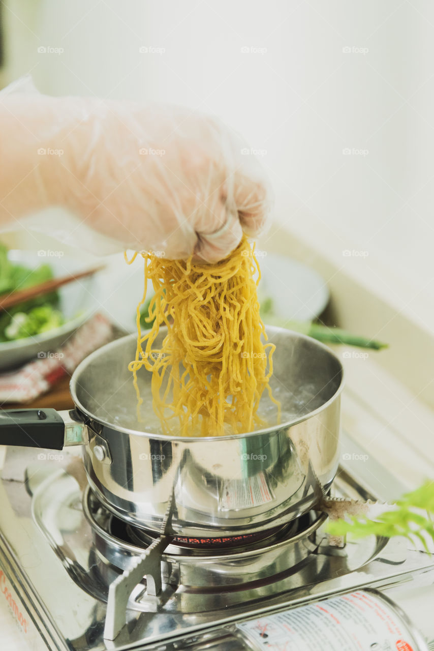 Noodle for lunch anyone? A woman’s hand in plastic glove, putting a noodle into a pot containing hot water, as a part of a process in making delicious noodle