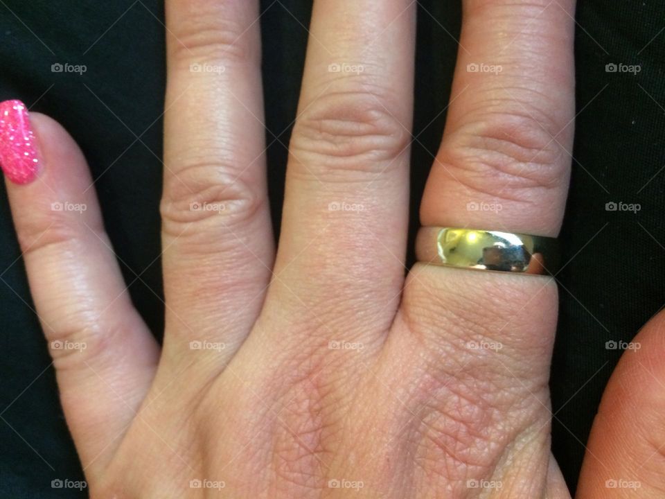 Ex’s 10 carat white gold wedding band worn on her middle aged left hand, on her pointer finger because they divorced, on a black background, and only her manicured pink painted gel nail of her pinky is showing