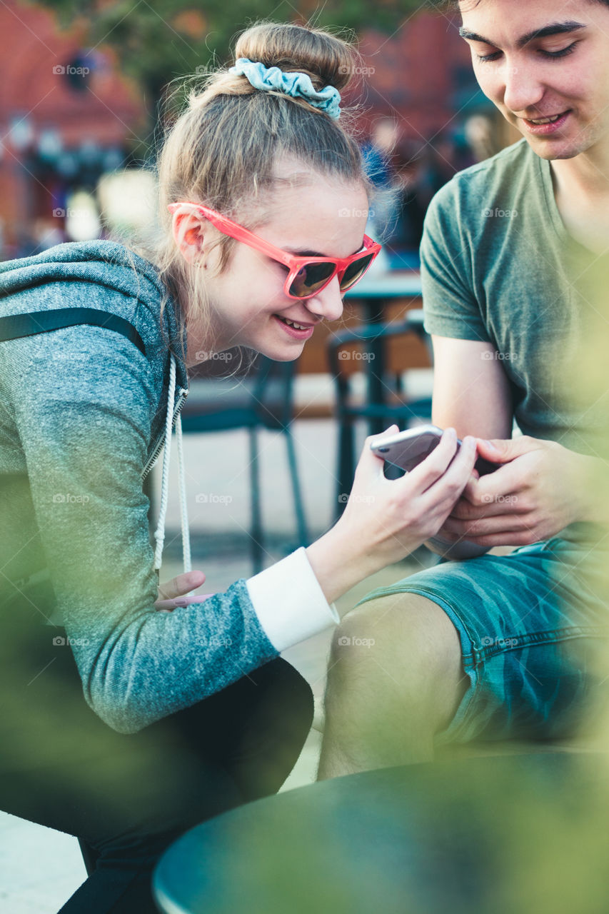 Couple of friends, teenage girl and boy, having fun with smartphones, sitting in center of town, spending time together