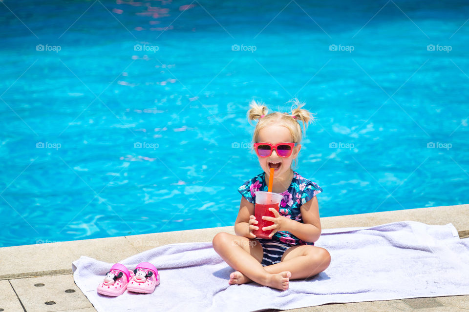 Cute little girl with blonde hair in sunglasses drinking fresh watermelon juice near swimming pool 