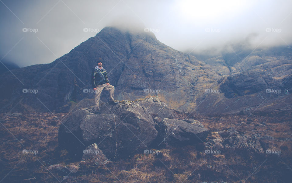 A man stands confidently upon some rocks, with large hills behind him, that are obscured with clouds in a mystical way