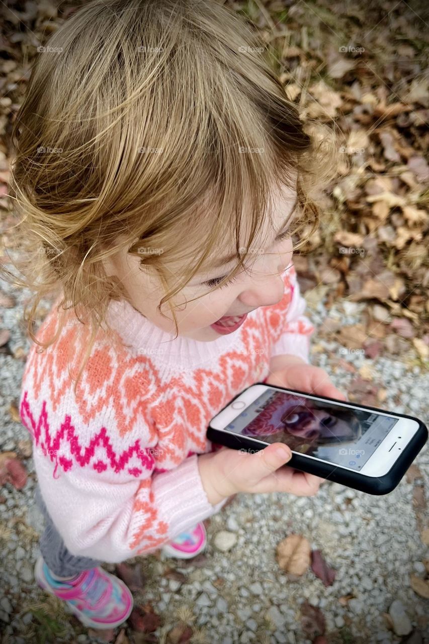 A young girl finds joy in seeing her picture on a phone as she plays outside on a crisp, Autumn day