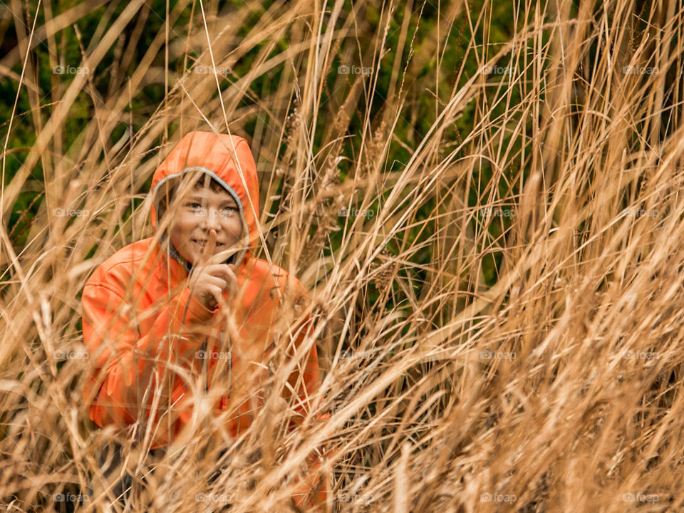 child in orange coat hiding in reeds with finger to mouth to stay quiet.