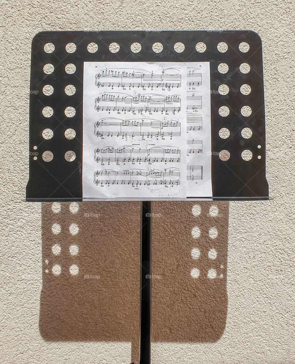 A black music stand on a leg with sheet music and notes casting a shadow on a sunny summer day against a textured wall