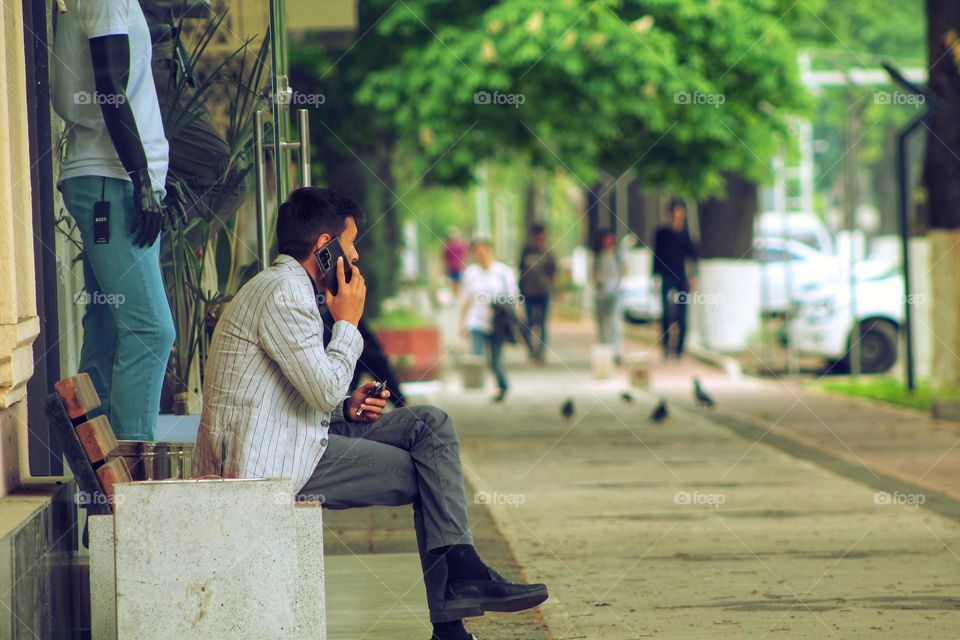 a young man in a white striped jacket sits on a street bench and talks on a mobile phone. Dot in the background are hurrying people and pigeons.