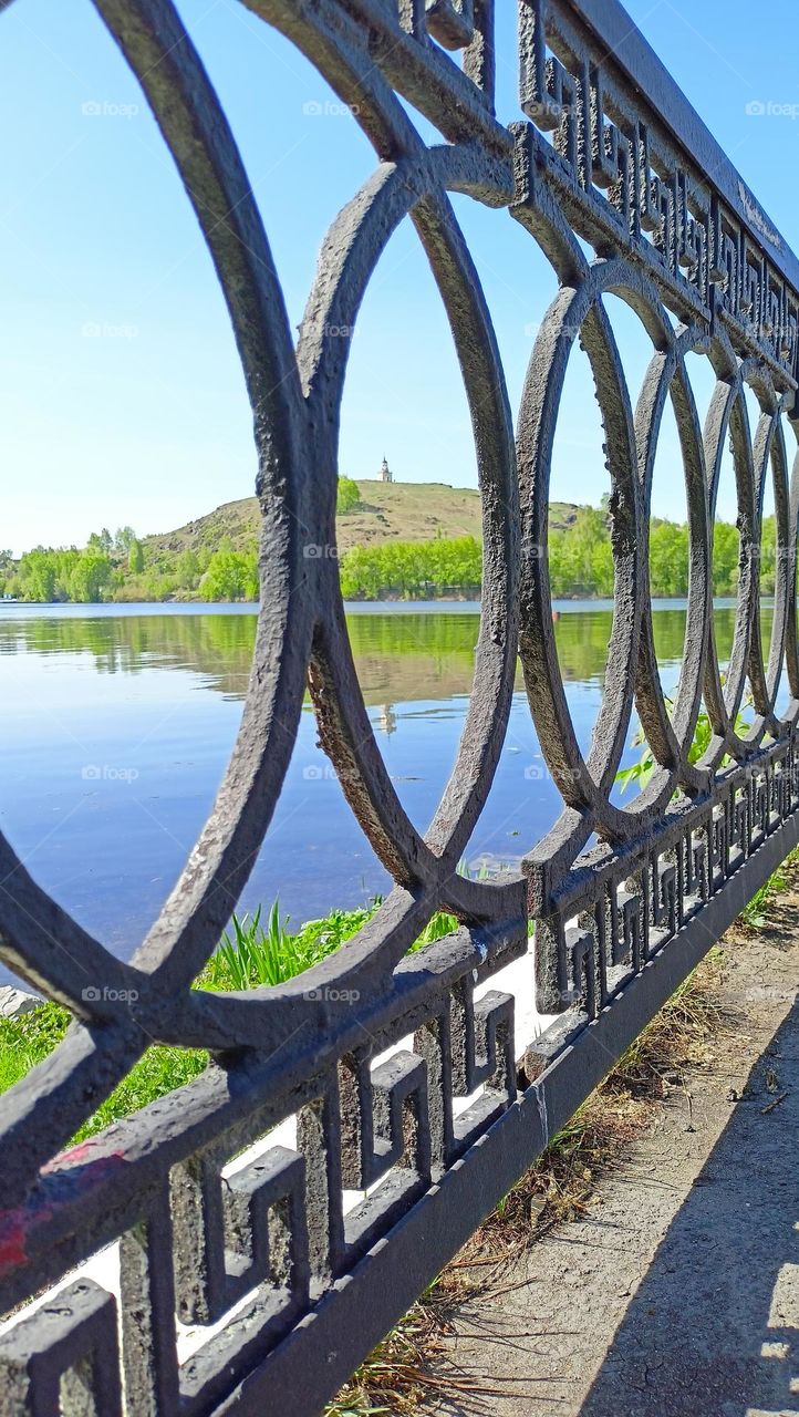 spring in the city, cityscape, pond, shore, mountain, buildings, architecture, iron fence, cloudy day, blue sky, water, park, view through