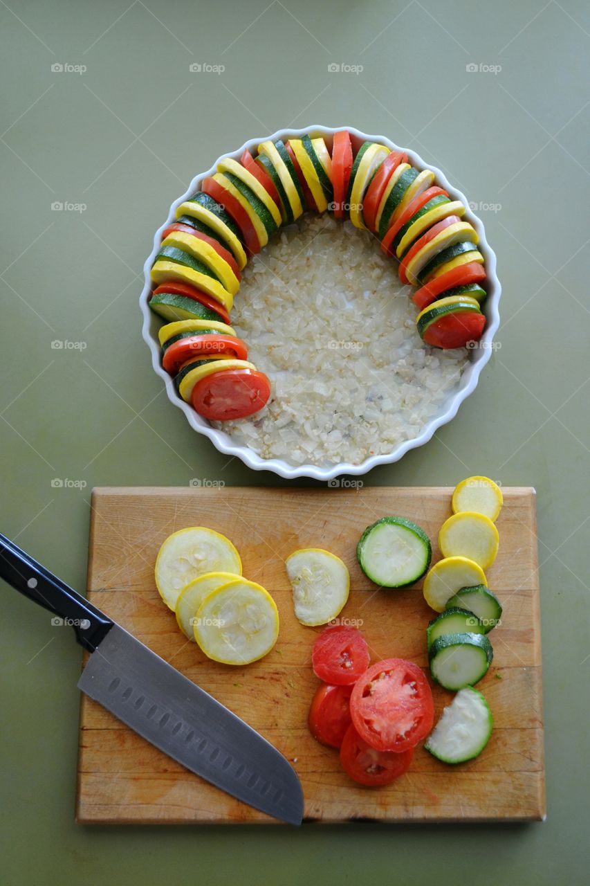 Vegetable Tian in process of being prepared with sliced tomatoes, zucchini, yellow or summer squash and potatoes on a bed on diced onions and garlic