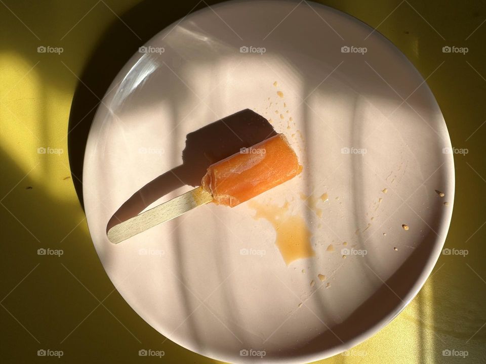 Half eaten orange popsicle on a tan plate atop a yellow metal bistro table with shadows