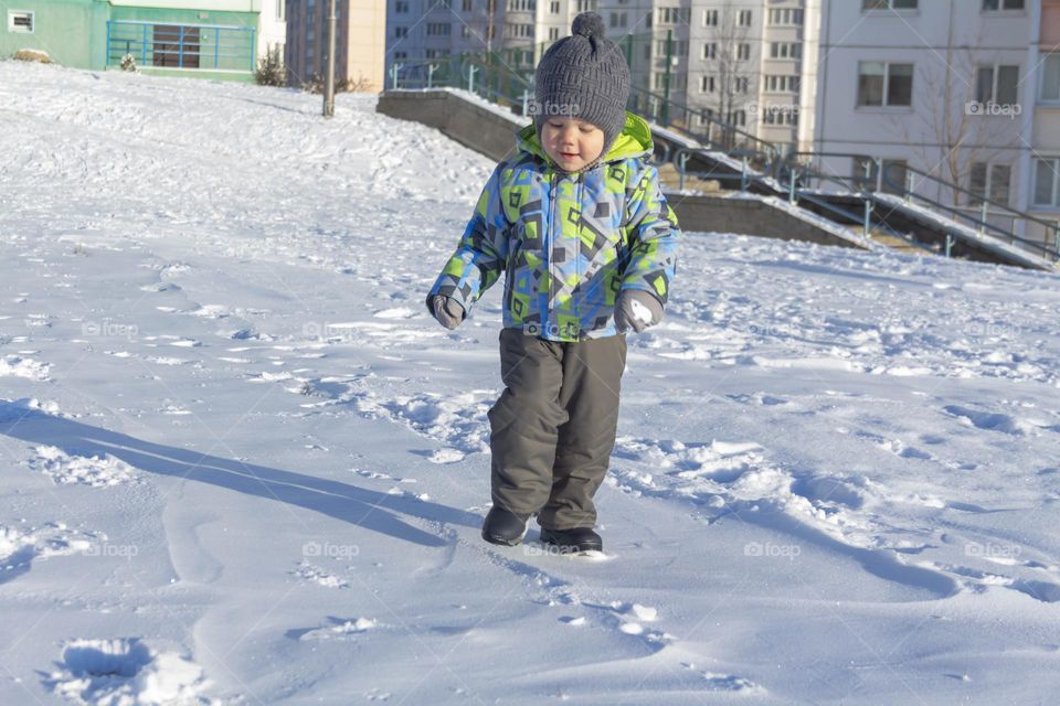 A child with a serious expression on his face in winter clothes jackets, pants, hat and boots in winter on the white snow on the street and in the park in nature plays winter fun.