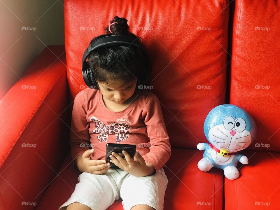 Baby girl listening music in phone by sitting on red couch beside her a blue toy 