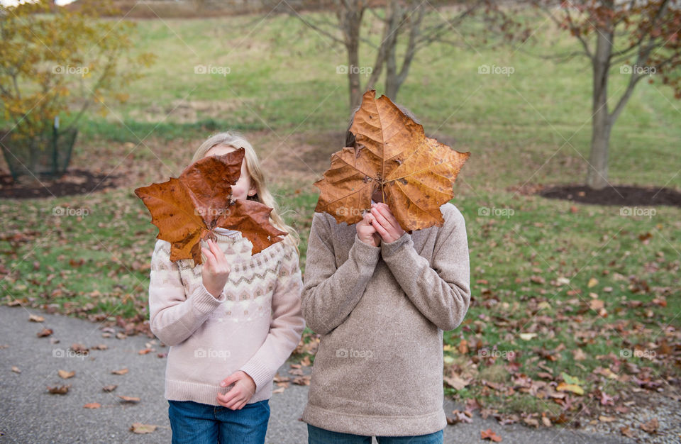Two children standing outdoors at a park during the fall and holding large maple leaves in front of their faces