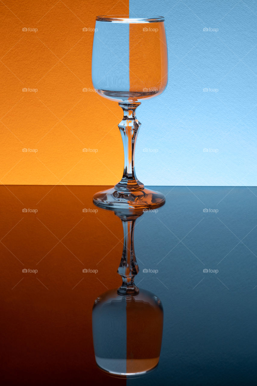 Glass of liquid on orange and blue background. Mirror table reflection