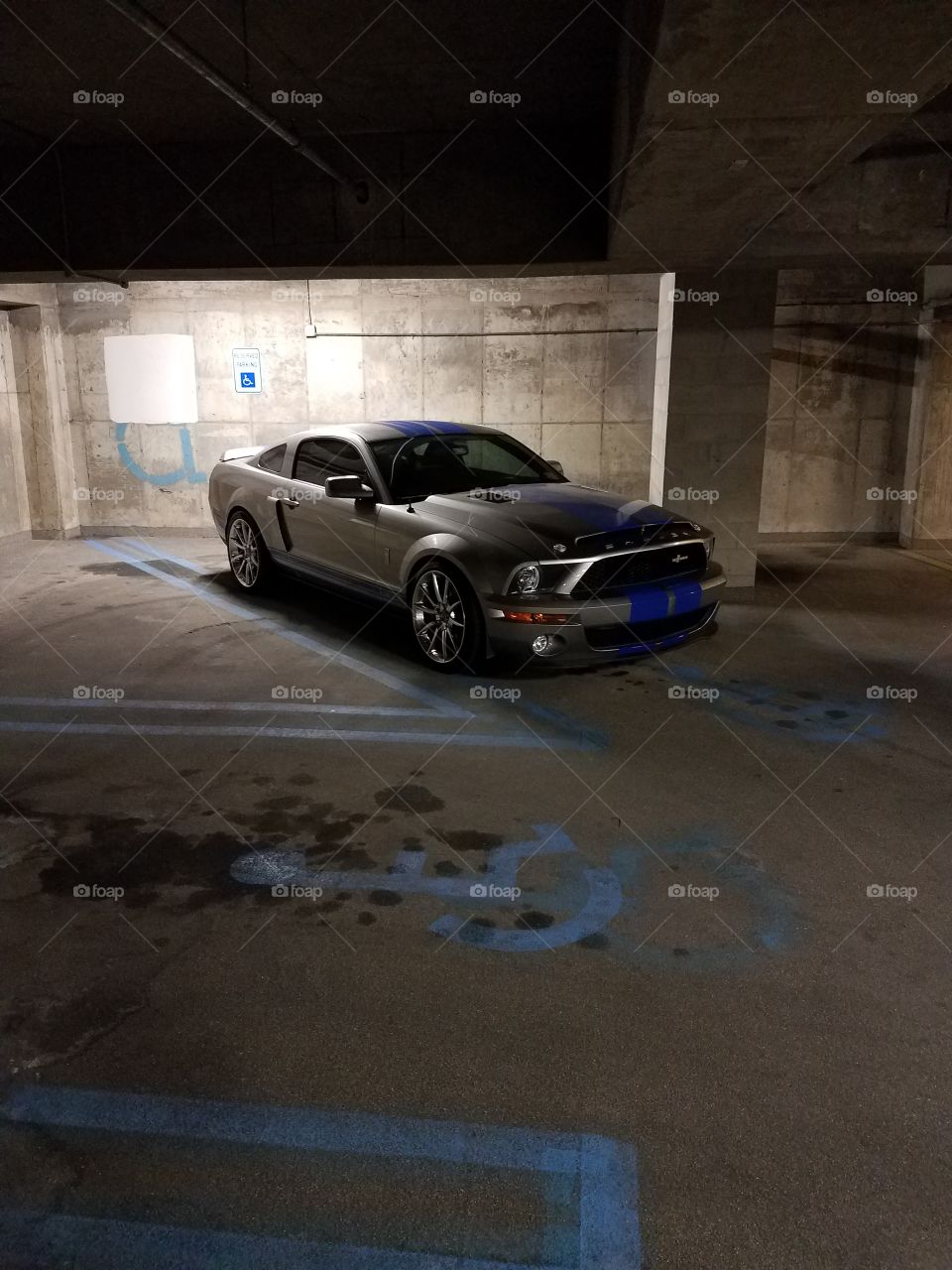 A silver Shelby mustang with blue racing stripes takes up a handicap spot in a garage.