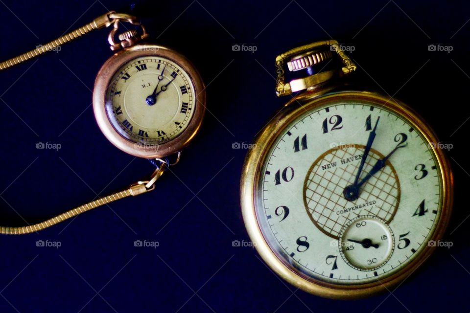 Flat lay of a woman’s gold vintage wristwatch with a slim expandable gold band and a man’s gold vintage pocket watch on a black surface
