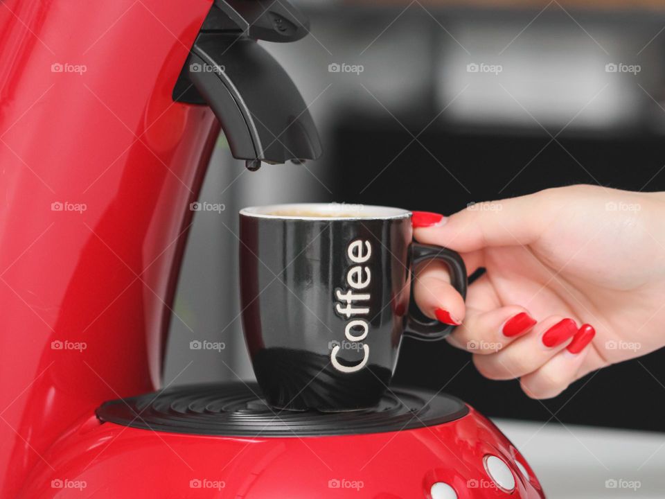 The hand of a young Caucasian girl with a beautiful short red manicure takes a mug of coffee from a coffee machine in the kitchen with a developed background,side view. The concept of a beautiful woman's manicure,morning breakfast,yusing technology.