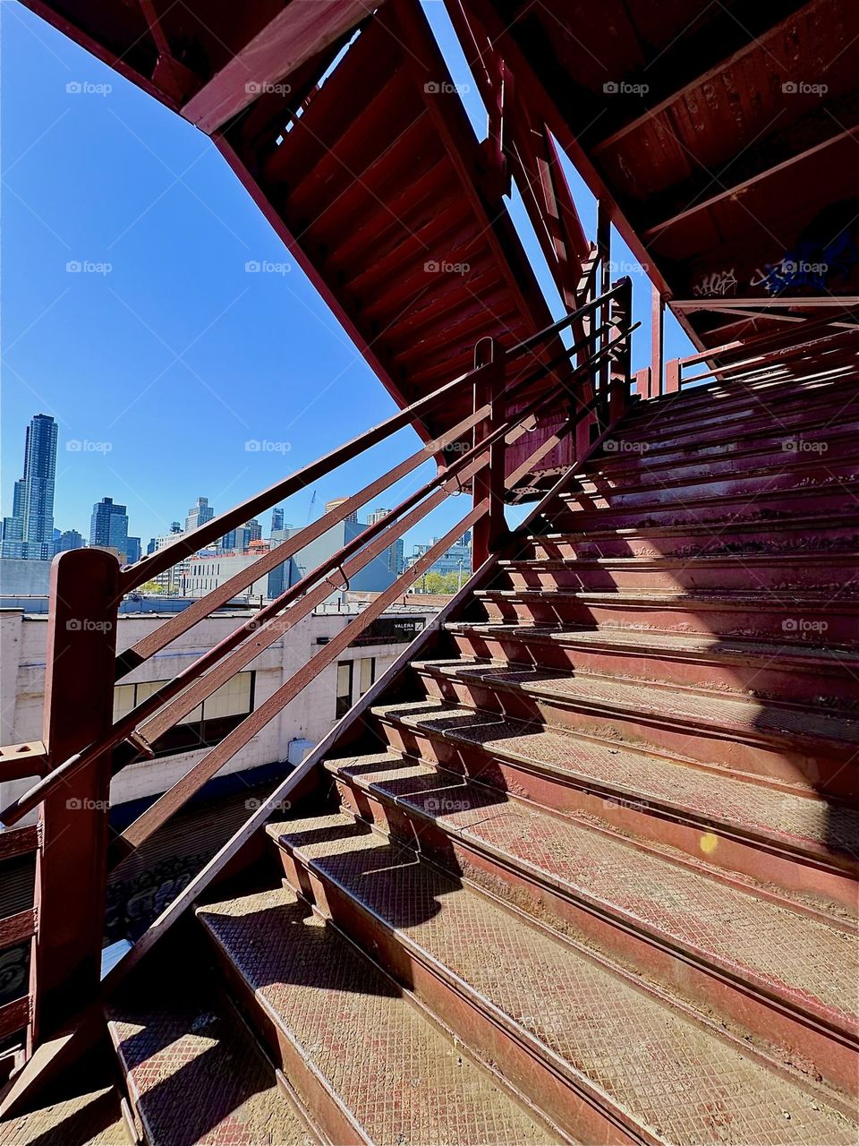 This is the red metal staircase of the “Pulaski Bridge” at “Newtown Creek” in LIC, Queens. The afternoon sun casts deep shadows that are displaced by the shape of the steps thus creating intricate patterns. 2024. Hypnotic Productions