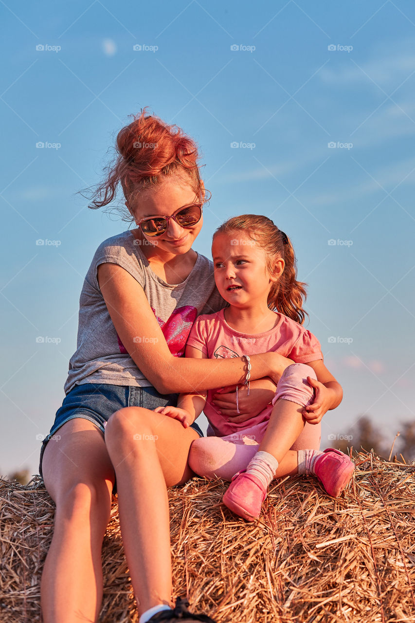 Mother and her daughters, teenage girl and her younger sister playing together on hay bale outdoors, spending vacations in the countryside. Candid people, real moments, authentic situations